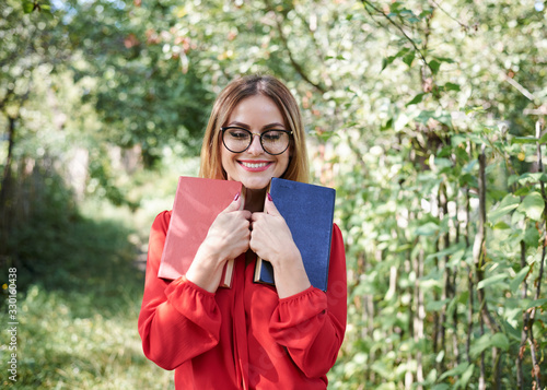 Young attractive blond woman in eyeglasses  wearing red shirt  holding red and dark blue books close to her face. Close-up portrait of pretty girl with her eyes closed. Student  dreaming of vacation.