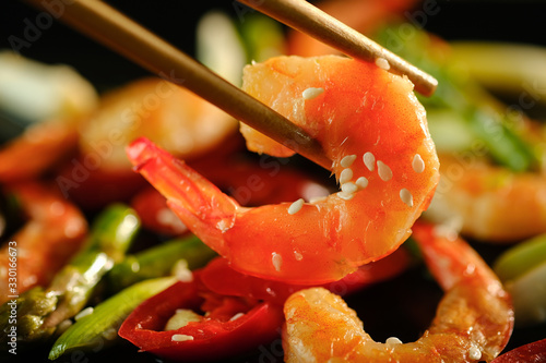 Shrimp is held with chopsticks on the black plate background with fried vegetables and shrimps.