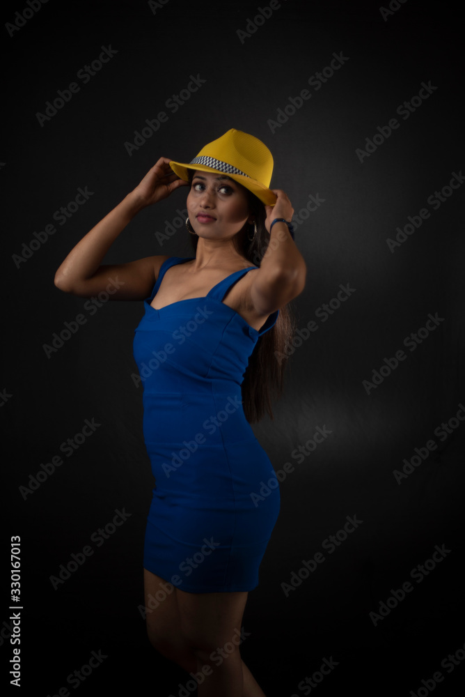 fashion portrait of an young and attractive Indian Bengali brunette girl with blue western dress and yellow cap in front of a black studio background. Indian fashion portrait and lifestyle. fashion portrait of an young and attractive Indian Bengali brunette girl with blue western dress and yellow cap in front of a black studio background. Indian fashion portrait and lifestyle.