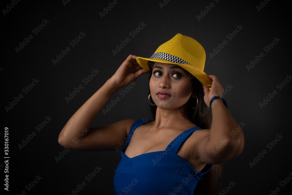 fashion portrait of an young and attractive Indian Bengali brunette girl with blue western dress and yellow cap in front of a black studio background. Indian fashion portrait and lifestyle. fashion portrait of an young and attractive Indian Bengali brunette girl with blue western dress and yellow cap in front of a black studio background. Indian fashion portrait and lifestyle.