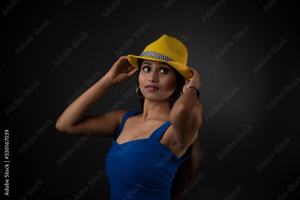 fashion portrait of an young and attractive Indian Bengali brunette girl with blue western dress, yellow cap and sunglass in front of a black studio background. Indian fashion portrait and lifestyle. fashion portrait of an young and attractive Indian Bengali brunette girl with blue western dress, yellow cap and sunglass in front of a black studio background. Indian fashion portrait and lifestyle.