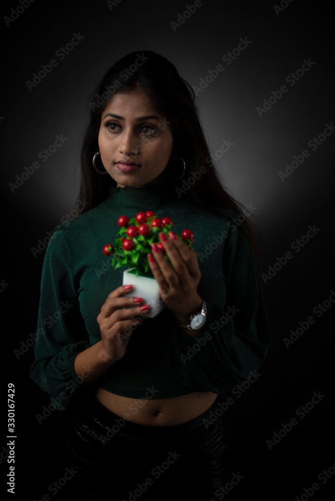 Fashion portrait of young and attractive Indian Bengali brunette girl with green cropped top and skirt holding cherry tree in front of black studio background. Indian fashion portrait and lifestyle. Fashion portrait of young and attractive Indian Bengali brunette girl with green cropped top and skirt holding cherry tree in front of black studio background. Indian fashion portrait and lifestyle.