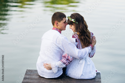 girl and a guy in embroidered clothing are sitting on a wooden pier and lean their heads. back view.