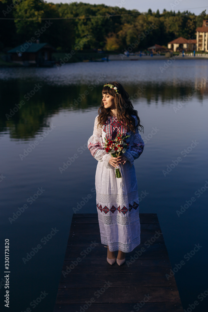 Fototapeta premium the bride in a chic embroidered dress stands on the pier against the background of the lake. girl holding a wedding bouquet.