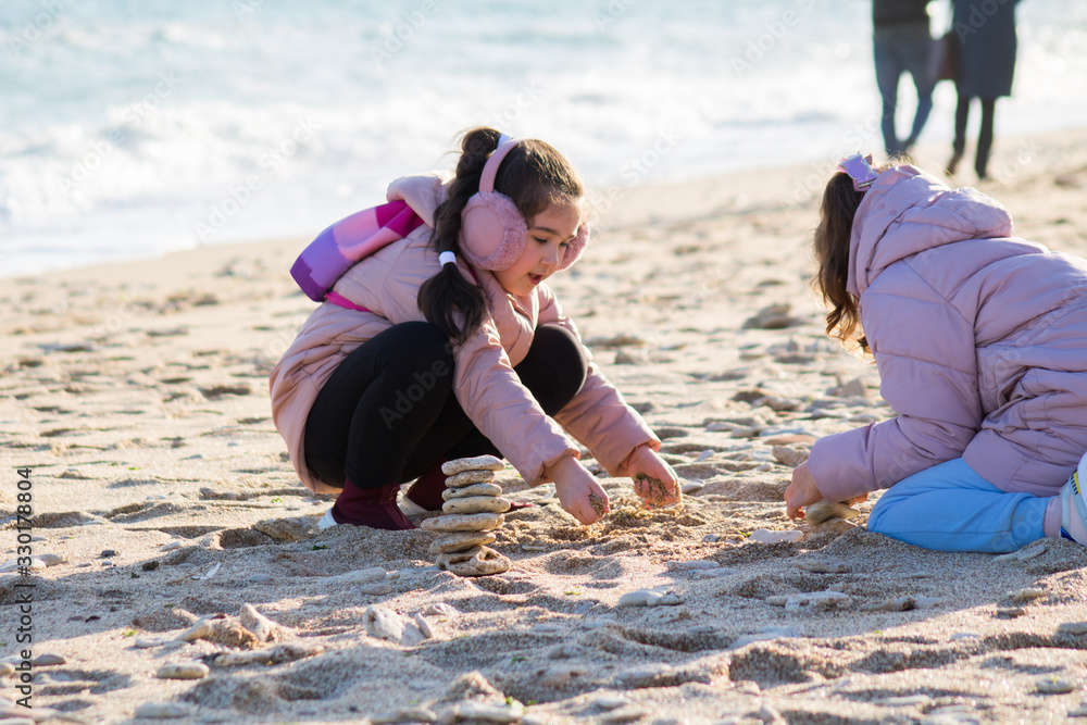 two little girls in pink jackets and earmuffs play in the sand on the beach, parents on the background two little girls in pink jackets and earmuffs play in the sand on the beach, parents on the background