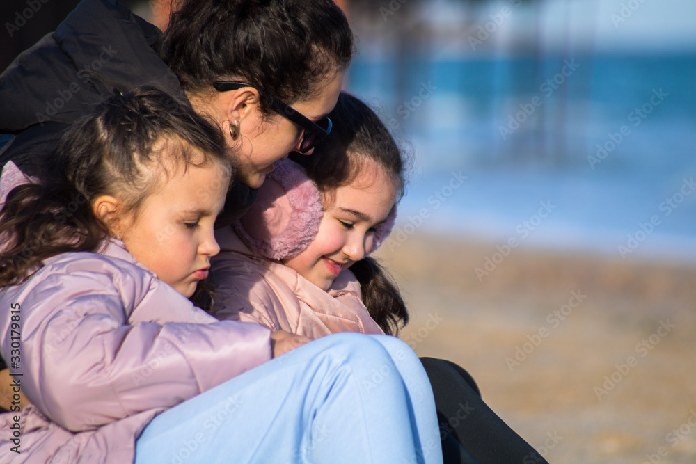 young mother in sunglasses hugs her little daughters in pink jackets on the beach