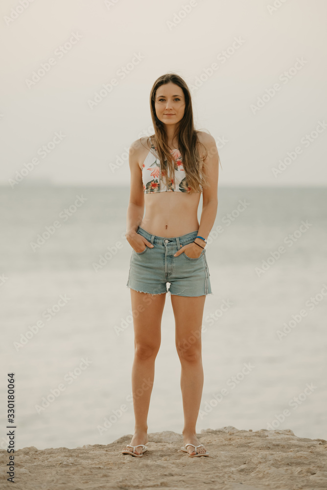 A pretty model with long hair in a swim top and denim shorts relaxing on the rocky shore of the red sea on the sunrise