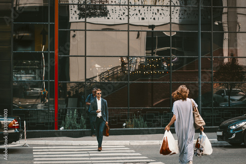 Successful young businessman dressed in stylish suit with smartphone and briefcase crossing zebra on metropolis street. Positive proud CEO concentraited on cellular strolling outdoors photo