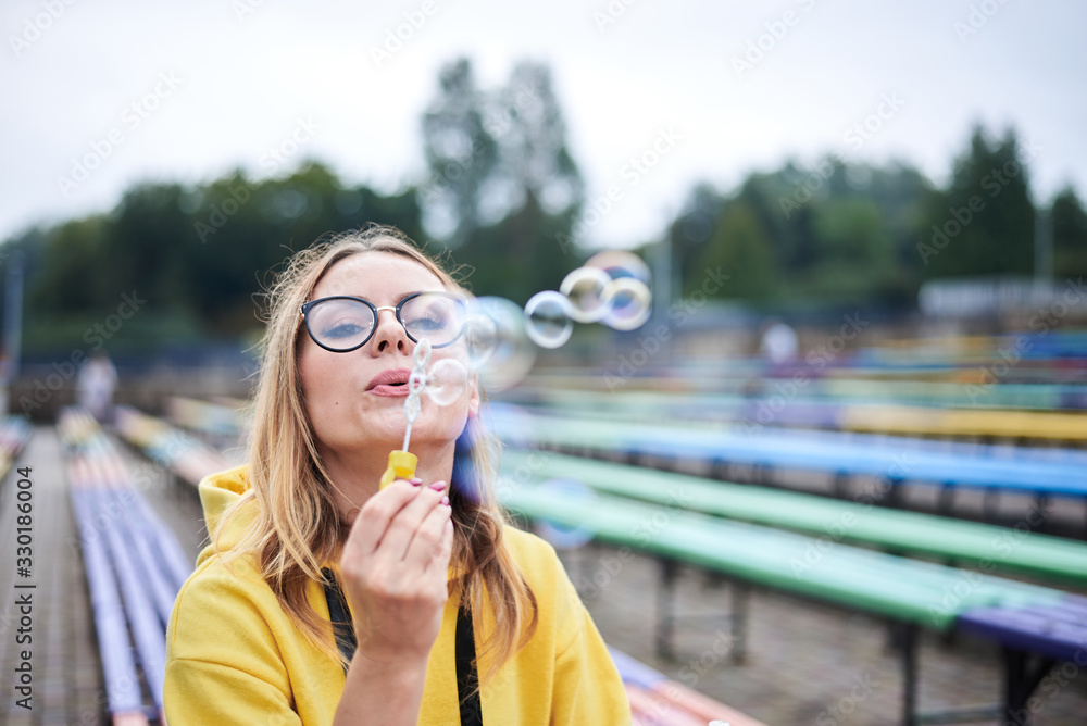 Young blond woman, wearing yellow hoody, blue jeans and eyeglasses, sitting on colorful bench in city urban park in summer. Close-up portrait of pretty girl, blowing making soap bubbles, laughing.