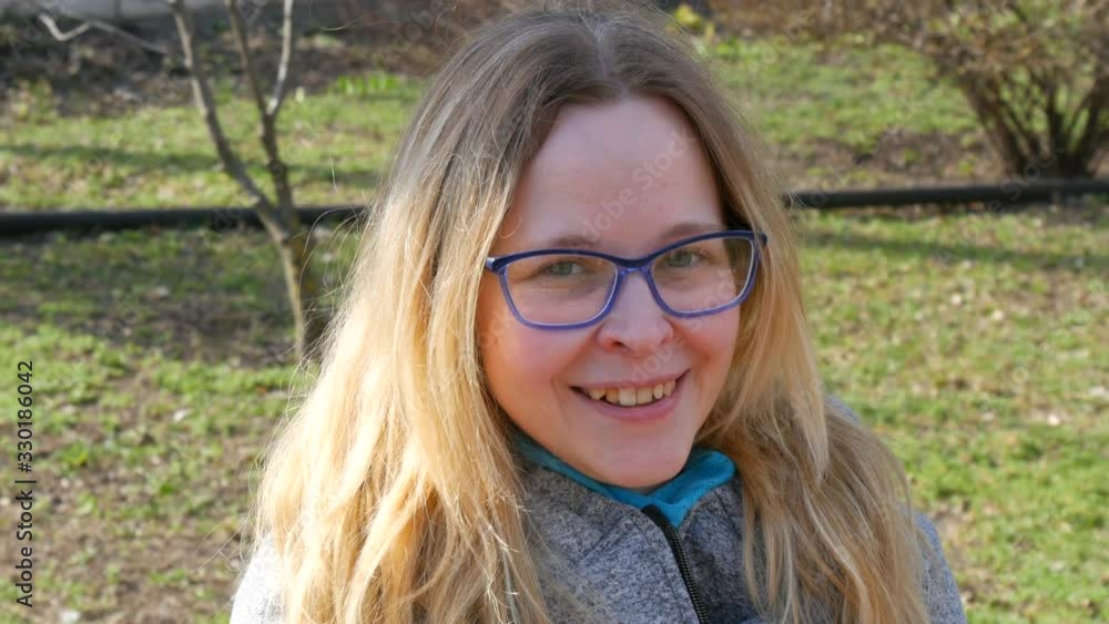 Beautiful young woman with blond hair and blue eyes in eyeglasses sits in a spring park and smiles. Beautiful young woman with blond hair and blue eyes in eyeglasses sits in a spring park and smiles.