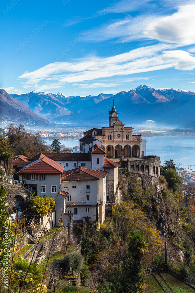 Stunning panorama view of Madonna del Sasso church above Locarno city on cliff with Lake Maggiore, snow covered Swiss Alps mountain peaks  blue sky cloud in background in autumn, Ticino, Switzerland