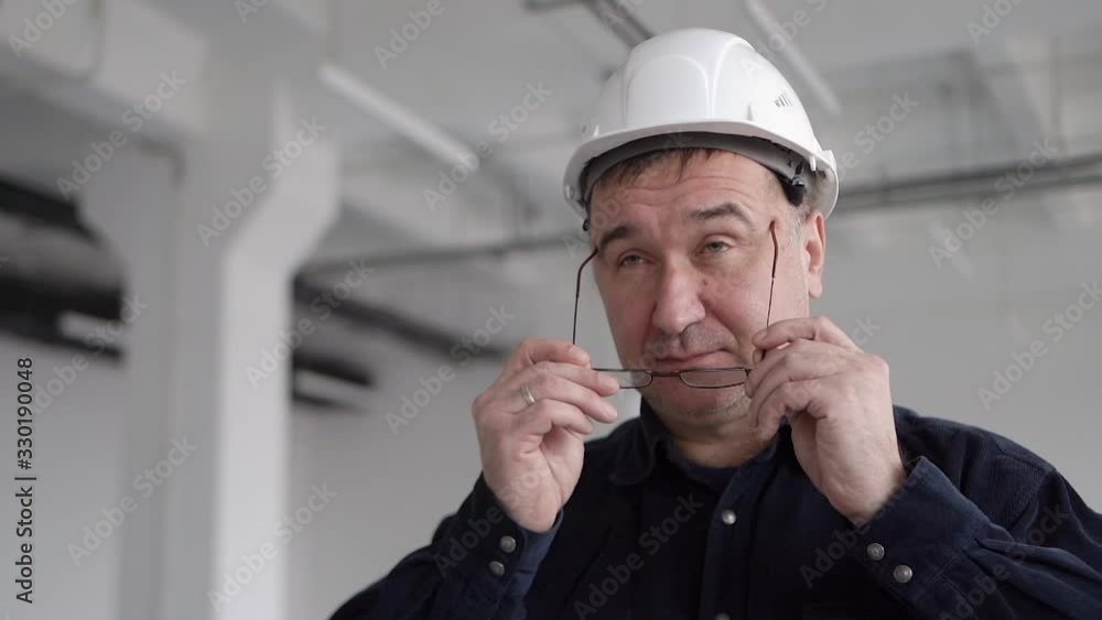An adult, a credible engineer, an architect in a blue shirt, stands in a large white room with columns, in a white protective helmet. He puts on glasses for vision and smiles at the camera An adult, a credible engineer, an architect in a blue shirt, stands in a large white room with columns, in a white protective helmet. He puts on glasses for vision and smiles at the camera