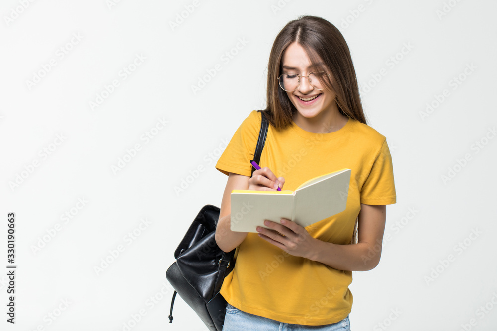 Portrait of a smiling casual girl student with backpack writing in a notepad while standing with books isolated over white background Portrait of a smiling casual girl student with backpack writing in a notepad while standing with books isolated over white background