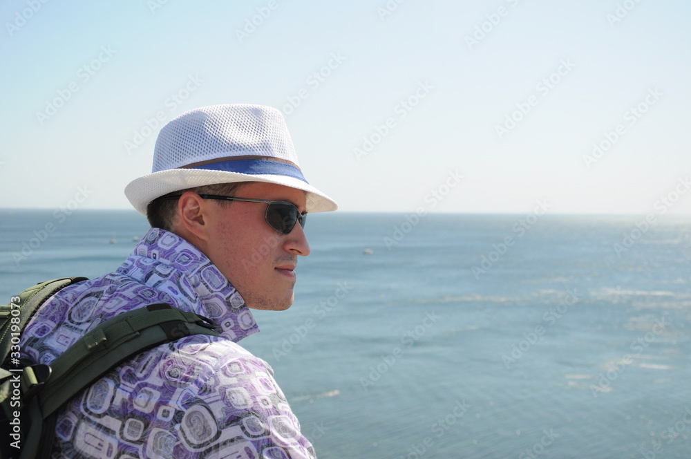 A young man in a white hat, sunglasses and a shirt with a raised collar looks away