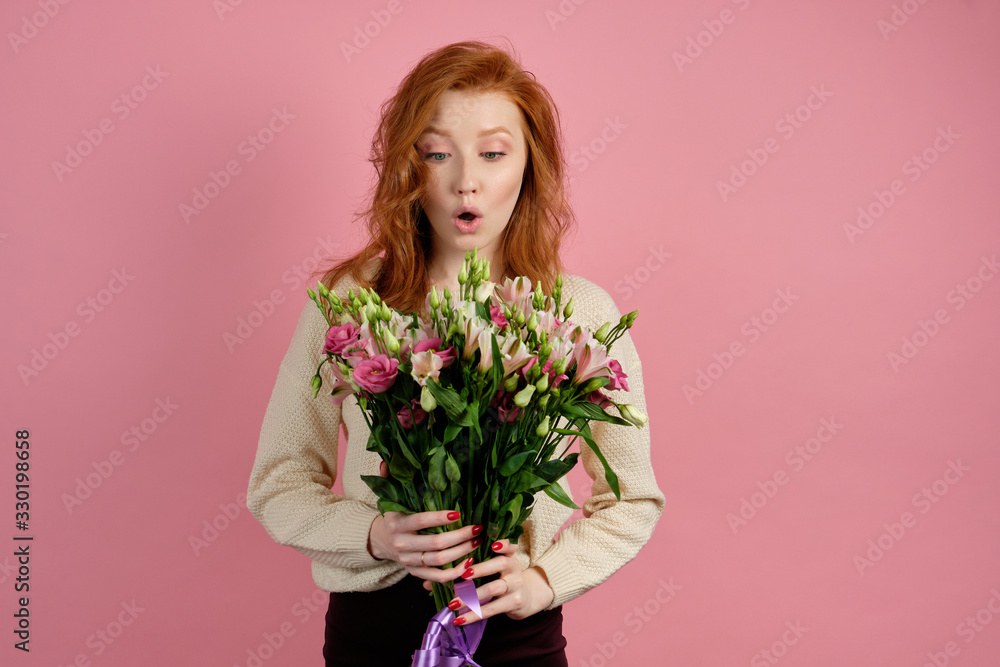 A beautiful red-haired girl stands on a pink background and is surprised to open her eyes and mouth looks at the bouquet in hands A beautiful red-haired girl stands on a pink background and is surprised to open her eyes and mouth looks at the bouquet in hands