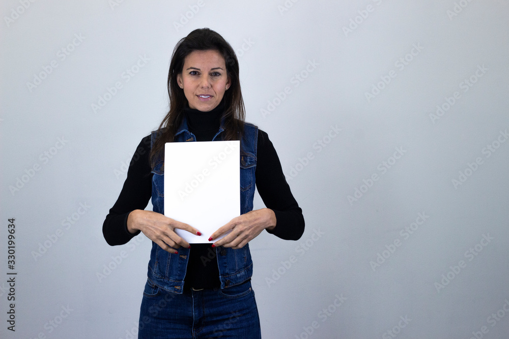 Portrait of attractive caucasian middle age woman holding white empty paper, isolated on gray background studio shot, black sweater, denim jacket, jeans. Place for your text in copy space.