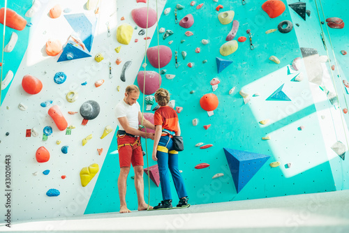Father helping his teenage son and checking climbing harness near the indoor climbing wall. Boy preparing.for a climbing. Happy parenting concept image.