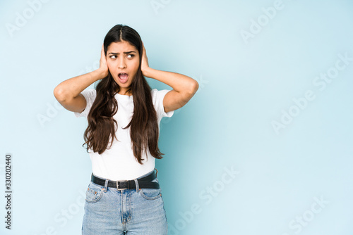 Young indian woman on blue background covering ears with hands trying not to hear too loud sound. © Asier