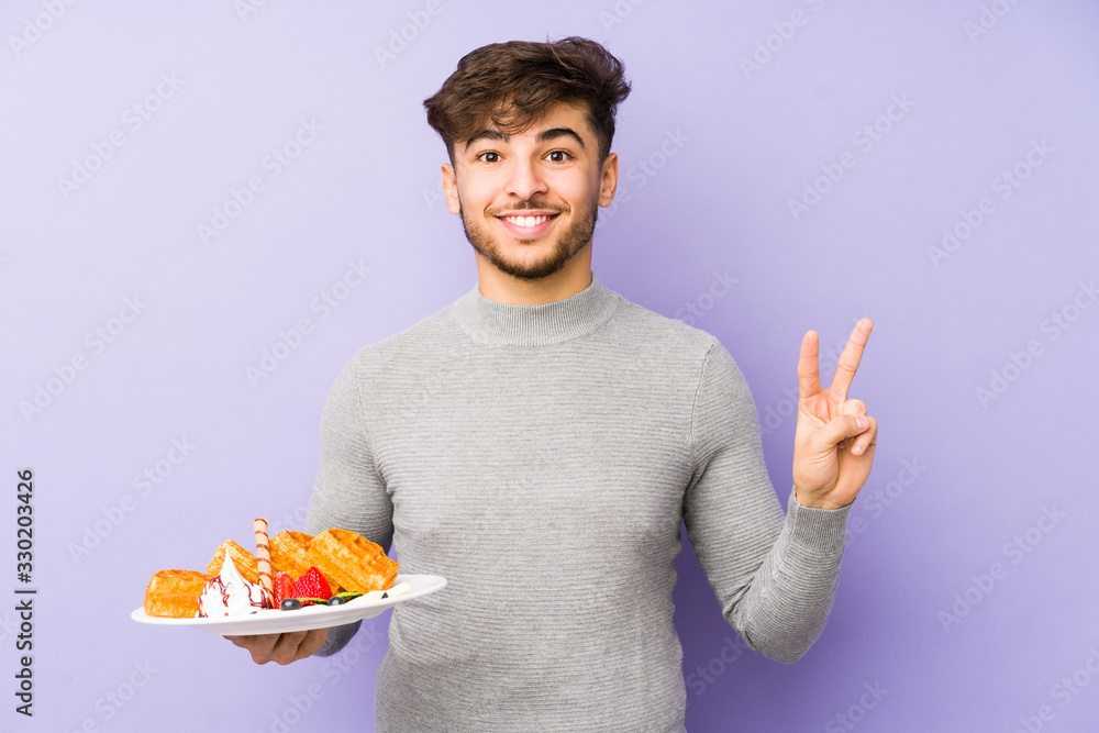 Young arabian man holding a waffle isolated joyful and carefree showing a peace symbol with fingers. Young arabian man holding a waffle isolated joyful and carefree showing a peace symbol with fingers.