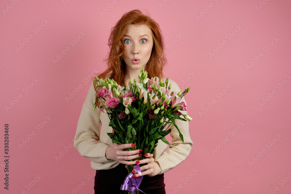 Beautiful red-haired girl holding a bouquet while standing on a pink background and opening her eyes wide and lips extended