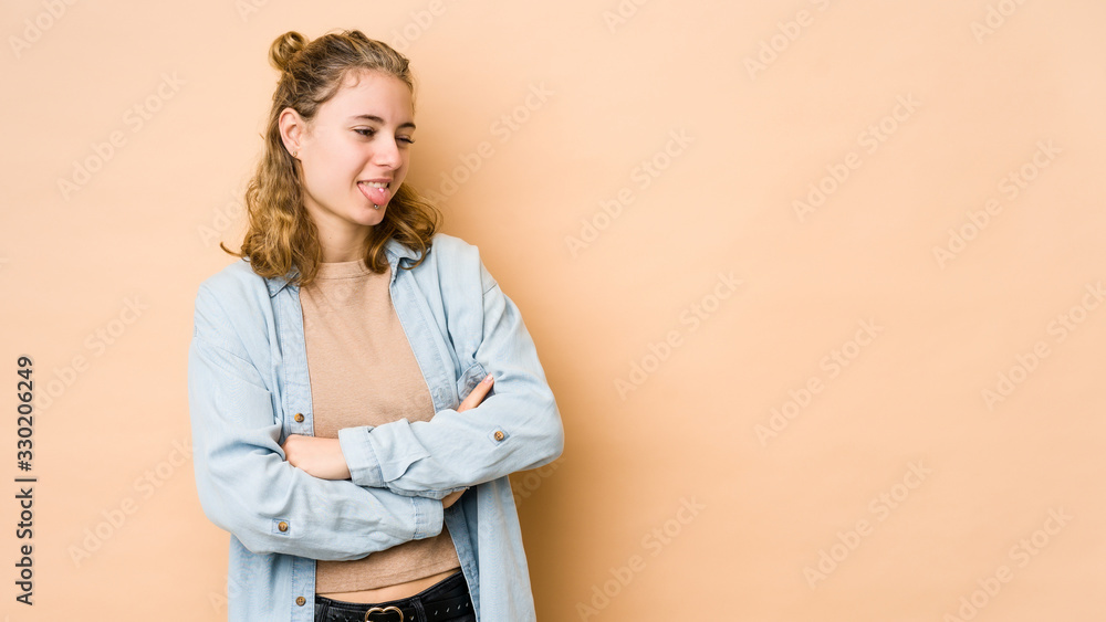 Young caucasian woman isolated on beige background funny and friendly sticking out tongue.