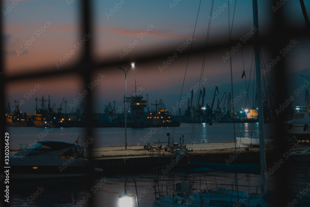 Stunning night view through a metal fence with a yacht and some ships for background.