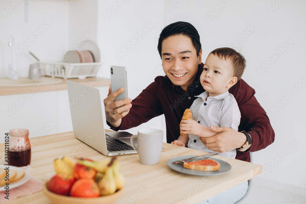 Asian dad and his little son having breakfast at laptop and taking selfie on smartphone Asian dad and his little son having breakfast at laptop and taking selfie on smartphone
