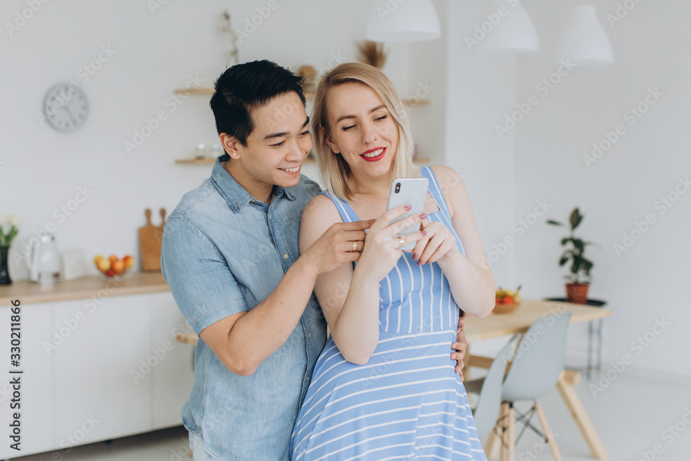 Asian man and his caucasian blonde woman spend time together in kitchen and looking at smartphone Asian man and his caucasian blonde woman spend time together in kitchen and looking at smartphone