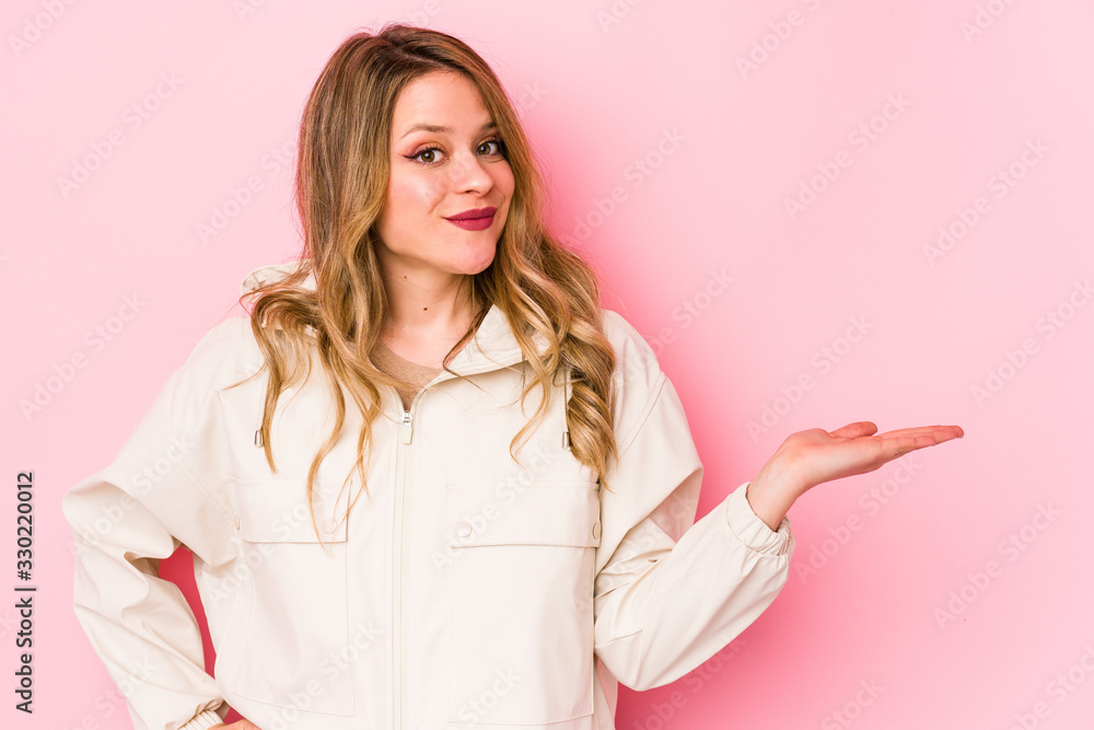 Young caucasian woman isolated on pink background showing a copy space on a palm and holding another hand on waist. Young caucasian woman isolated on pink background showing a copy space on a palm and holding another hand on waist.