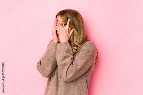 Young caucasian woman isolated on pink background blink through fingers frightened and nervous.