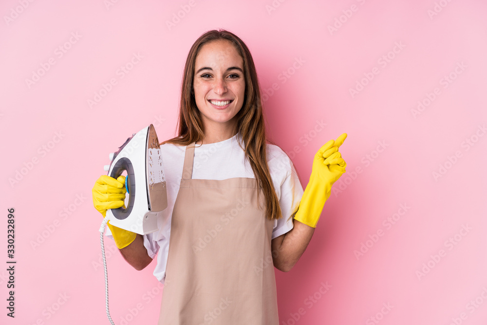 Young caucasian woman with clothes iron isolated smiling and pointing aside, showing something at blank space. Young caucasian woman with clothes iron isolated smiling and pointing aside, showing something at blank space.
