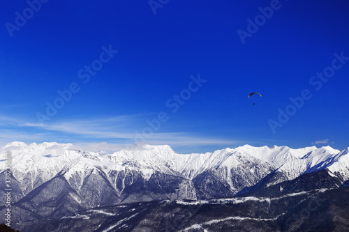 Winter peaks of Caucasus mountain range and blue sky with flying paraglider. Rosa Khotor ski resort, Russia. High mountains covered with snow.