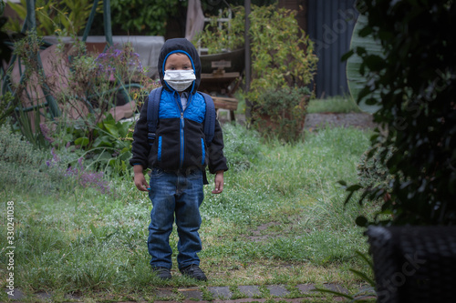A young boy wearing a backpack, face mask, hoodie jacket, and rain boots, standing outdoors while its raining.