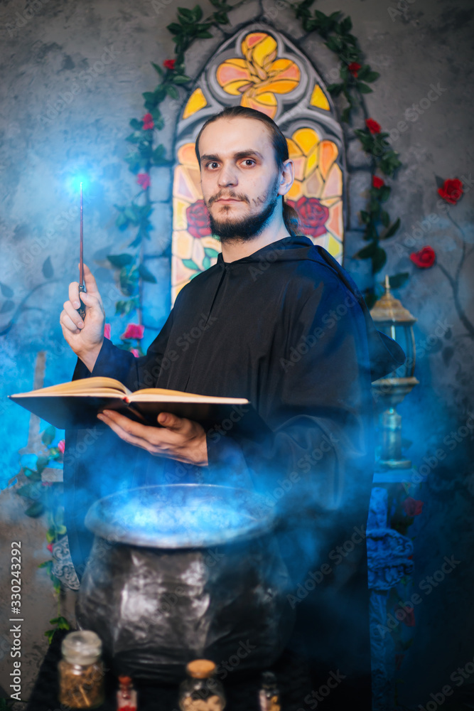 Fototapeta premium The mage is cooking a potion. A male wizard in a black mantle with a magic wand and a book. He casts a spell over the witch's cauldron. Guy in an old castle with luminous magic