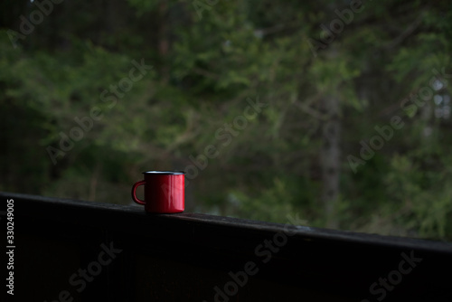 a red coffee mug stands on the railing of the wooden verandah, steam escapes from it, and green pines stand against the background