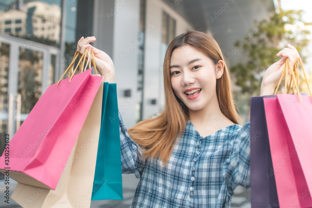 Smiling young Asian woman with shopping colour bags over mall background. using a smart phone shopping online and smiling while standing mall building. lifestyle concept Smiling young Asian woman with shopping colour bags over mall background. using a smart phone shopping online and smiling while standing mall building. lifestyle concept