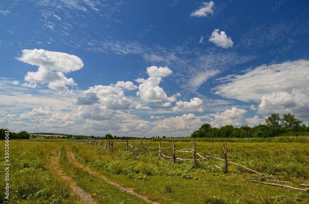 landscape with a country road, cattle fence, field, forest, and clouds at summer