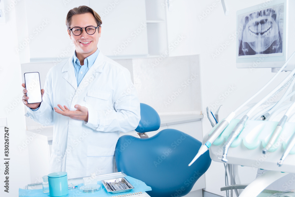 Portrait of smiling man in white doctor robe uniform, glasses is holding smartphone with blank screen, inviting patient to visit, standing in office, clinic. Dentist workplace. Dentistry concept. Portrait of smiling man in white doctor robe uniform, glasses is holding smartphone with blank screen, inviting patient to visit, standing in office, clinic. Dentist workplace. Dentistry concept.