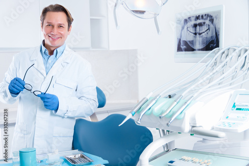 Portrait of smiling man in white doctor robe uniform is standing in office, clinic with chair, instruments. Dentist workplace. Orthodontist is preparing for visit of patient. Dentistry concept.