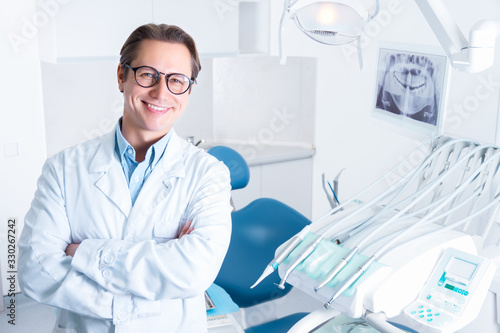 Portrait of smiling man in white doctor robe uniform is standing in office  clinic with chair  instruments. Dentist workplace. Orthodontist is preparing for visit of patient. Dentistry concept.