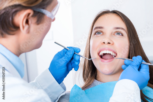 Smiling young woman is sitting in dental blue chair in clinic  office. Man doctor  orthodontist is conducting examination  treating teeth with tools  instruments. Visit to dentist concept.