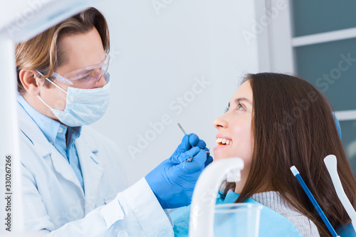 Smiling young woman is sitting in dental blue chair in clinic  office. Man doctor  orthodontist is conducting examination  treating teeth with tools  instruments. Visit to dentist concept.