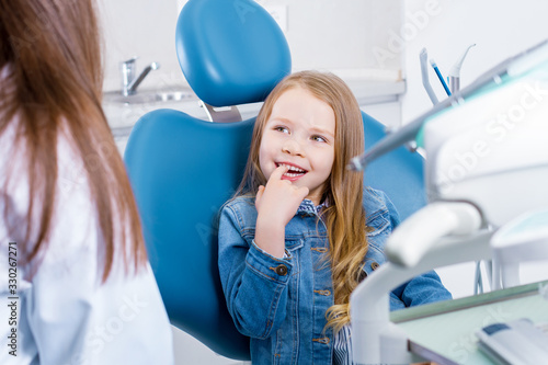 Little cute smiling girl is sitting in blue dental chair in clinic, office. Woman doctor is preparing for examination of child teeth, talking with patient. Visiting dentist with children.