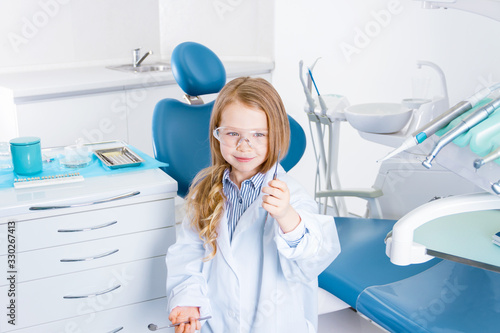 Little cute girl in white doctors robe uniform and protective glasses is standing in front of chair in office with instruments and tools. Child is playing in dentist, orthodontist. Dentistry concept.