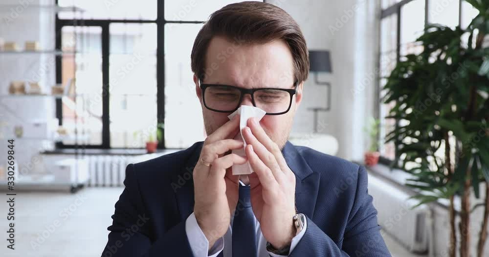 Sick allergic businessman having allergy blowing running nose at workplace. Ill man executive wearing suit feeling unhealthy suffering from seasonal allergic rhinitis or flu symptom in office concept. Sick allergic businessman having allergy blowing running nose at workplace. Ill man executive wearing suit feeling unhealthy suffering from seasonal allergic rhinitis or flu symptom in office concept.
