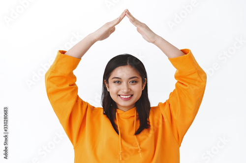 Girl fill secured. Close-up portrait cute silly asian woman in orange hoodie making roof with hands above head smiling joyfully, concept of safeness, family and reliability, stand white background © Cookie Studio