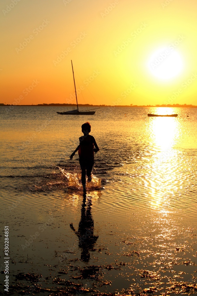 silhouette of child running on the seashore at sunset with the headland in the background and boats in the distance