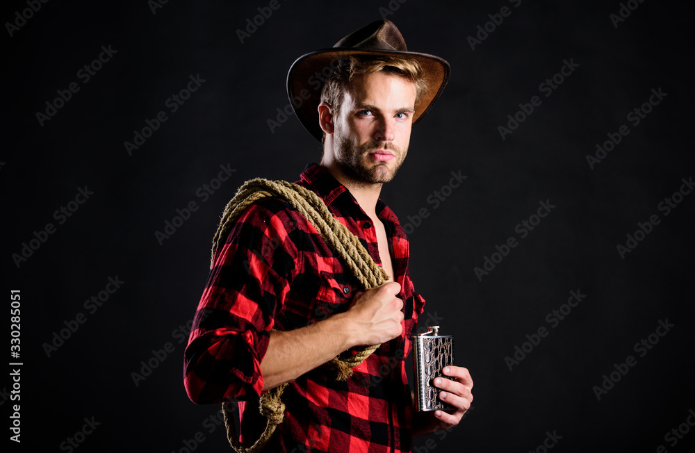 Fototapeta premium Brutal cowboy drinking alcohol. Western culture. Man handsome unshaven cowboy black background. Western life. Man wearing hat hold rope and flask. Lasso tool of American cowboy. Sheriff concept