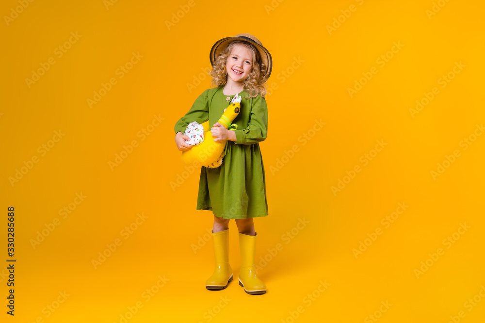 little girl smiles in a green linen dress, yellow rubber boots and a straw hat holding a textile toy chicken on a yellow background of isolate. concept of eco-friendly food and farming little girl smiles in a green linen dress, yellow rubber boots and a straw hat holding a textile toy chicken on a yellow background of isolate. concept of eco-friendly food and farming