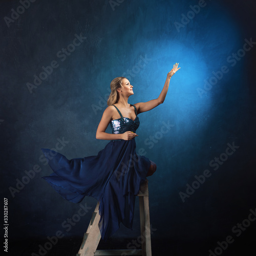 girl on the ladder reaches up with her hands. A young woman in a blue dress on a blue background